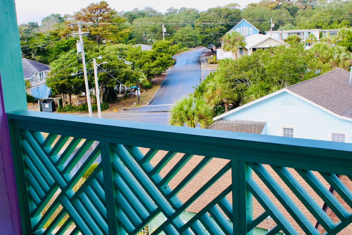 A relaxed view from the balcony shows the surrounding trees and houses across the street, with a glimpse of the tranquil environment just outside the property. The scene suggests leisurely mornings and easy walks to explore the neighborhood or head straight to the beach.