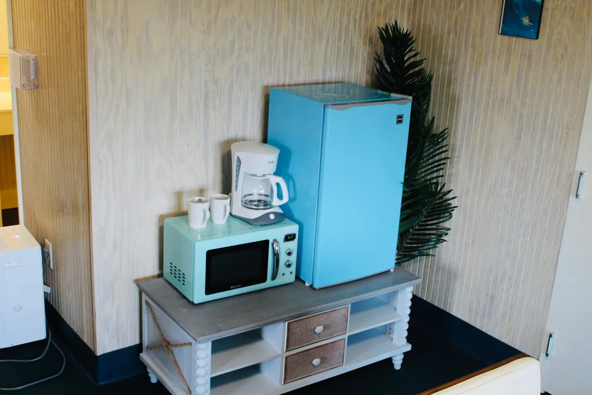 A cozy, light-filled kitchenette area featuring a light blue refrigerator, a modern coffee maker, a microwave, and a small table with drawers. A green plant adds a touch of freshness to the inviting setup, and all appliances are within easy reach, showcasing efficient use of space.
