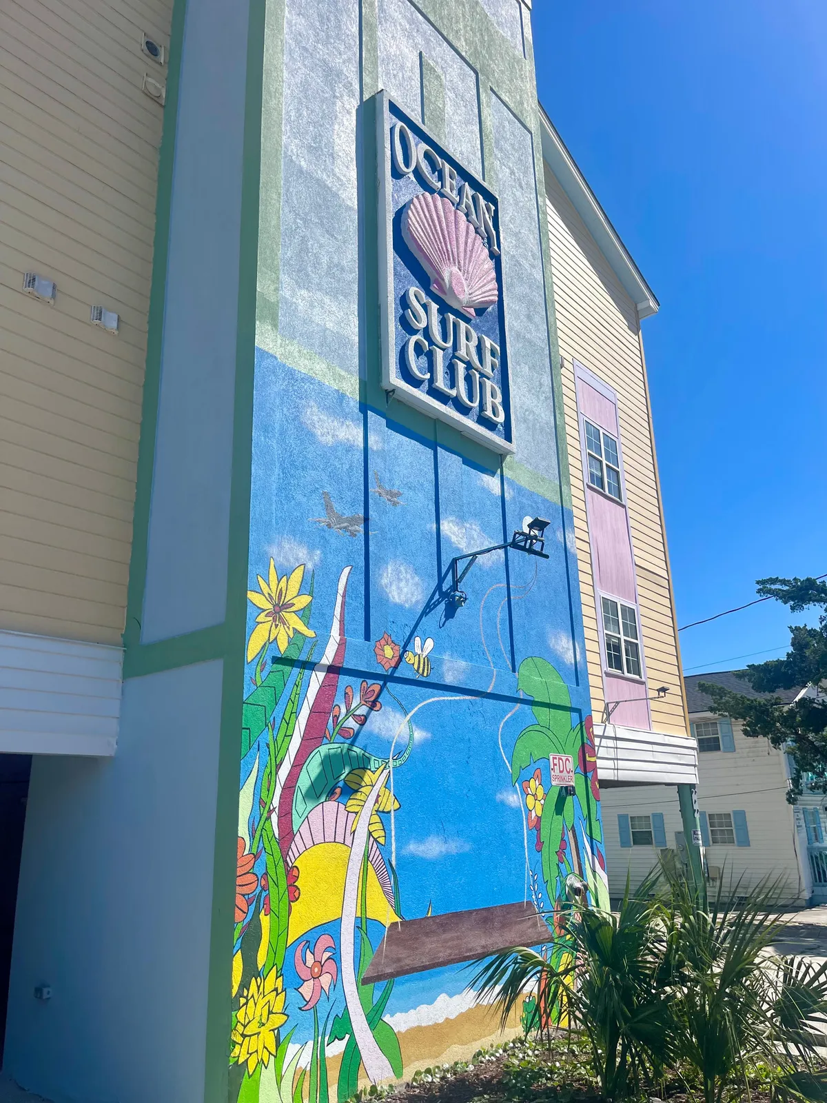 This exterior image captures the vibrant Ocean Surf Club sign set against a colorful mural, with a swing in the foreground. The scene radiates a playful, inviting atmosphere that hints at the community's fun-loving spirit. The setting sun bathes the area in warm light, making the entrance feel welcoming and full of energy.