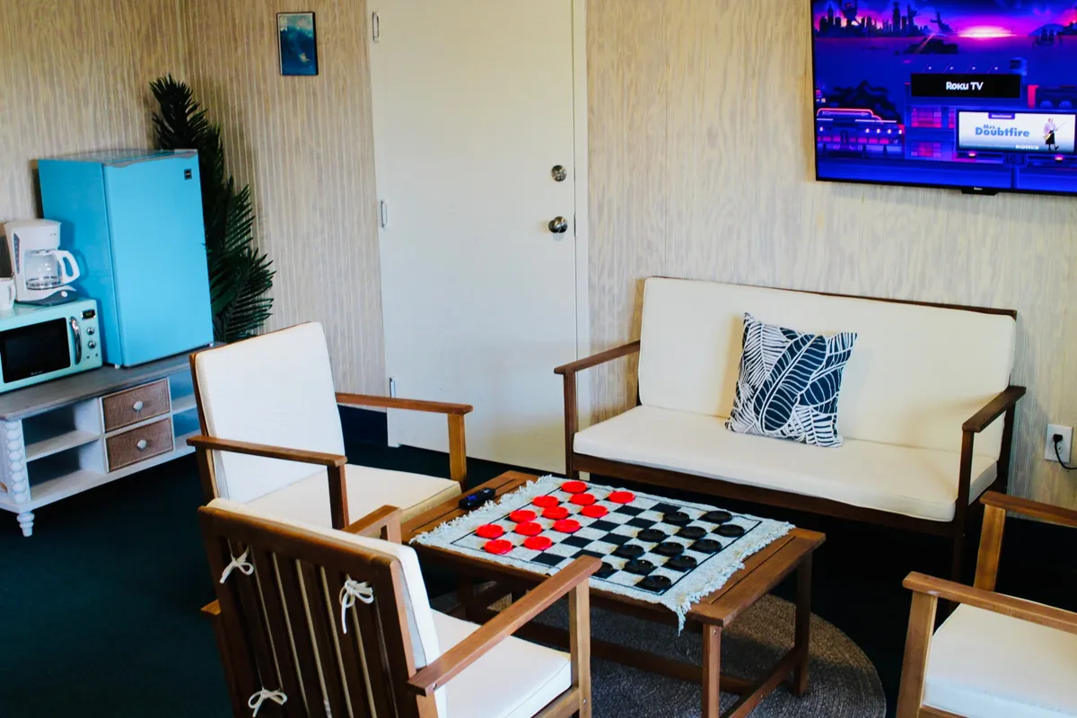 A cozy living area featuring a sofa with a decorative cushion, a blue refrigerator, and a checkerboard game set up on a table. The space exudes a casual, friendly vibe, ideal for socializing, relaxing, or enjoying family game nights.