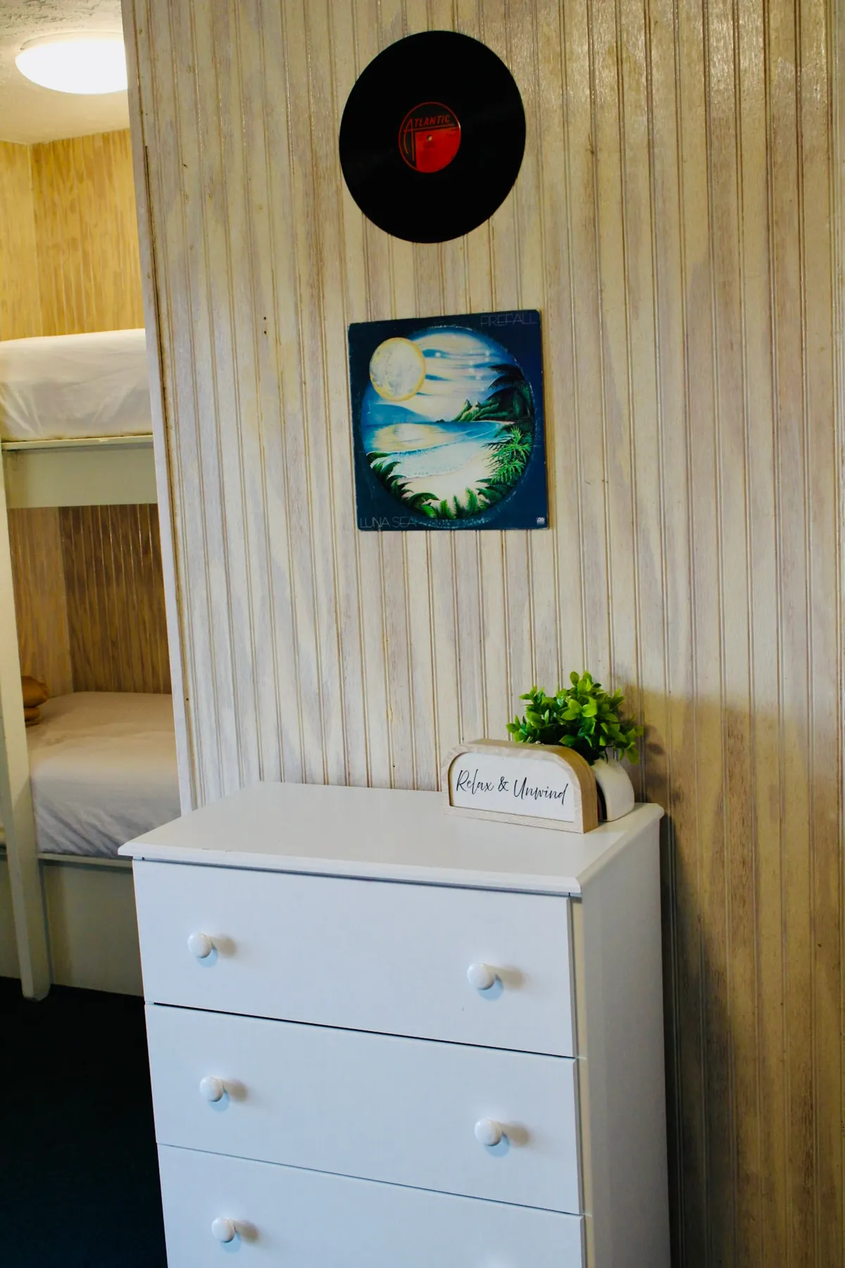 A thoughtfully decorated corner featuring a white dresser, decorative vinyl record, artwork, a small plant, and an uplifting sign on the wall. The decor is playful yet calming, adding warmth and personal style to the living space.